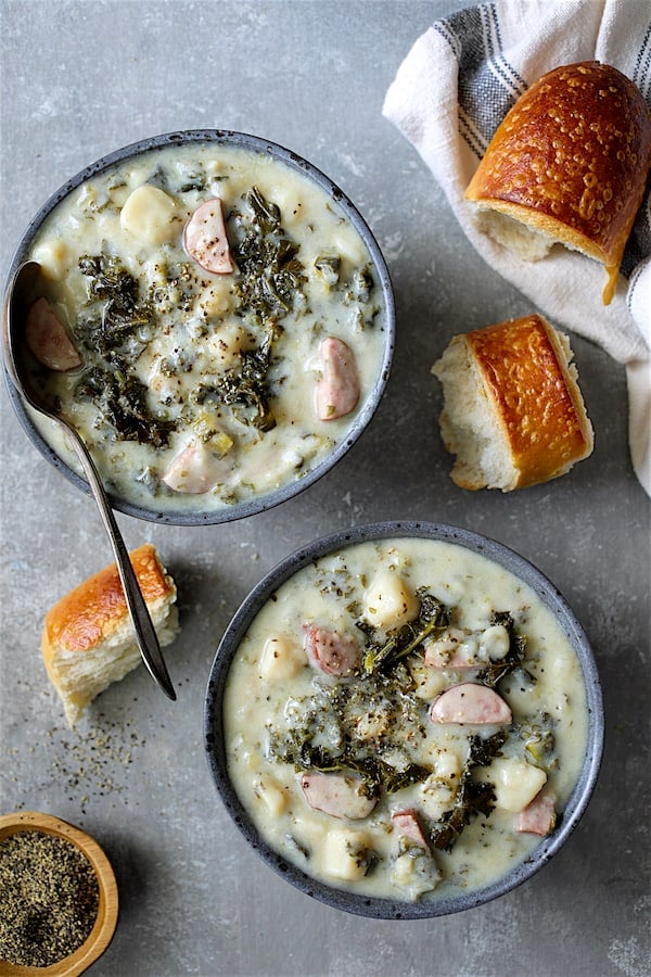 Photo of two servings of Creamy Potato Kielbasa and Kale Soup in gray bowls with bread scattered about.