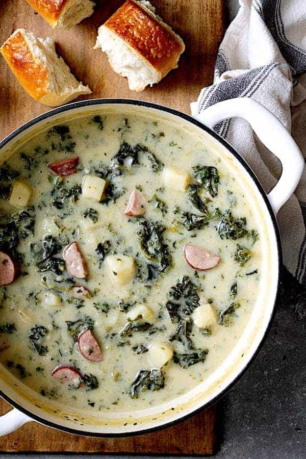Photo of Creamy Potato Kielbasa and Kale Soup in white Dutch oven with bread on a cutting board.