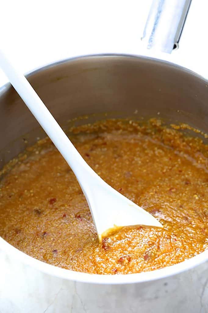 Photo of pumpkin grits in stainless steel saucepan being stirred with white spoon.