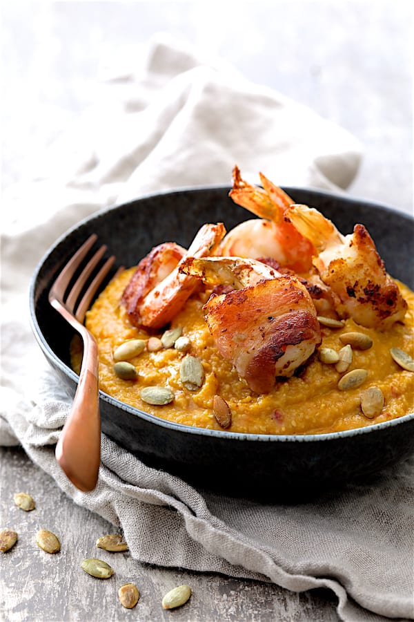Photo of Pumpkin Chipotle and White Cheddar Grits with Bacon Wrapped Shrimp in black bowl on gray napkin garnished with pumpkin seeds.