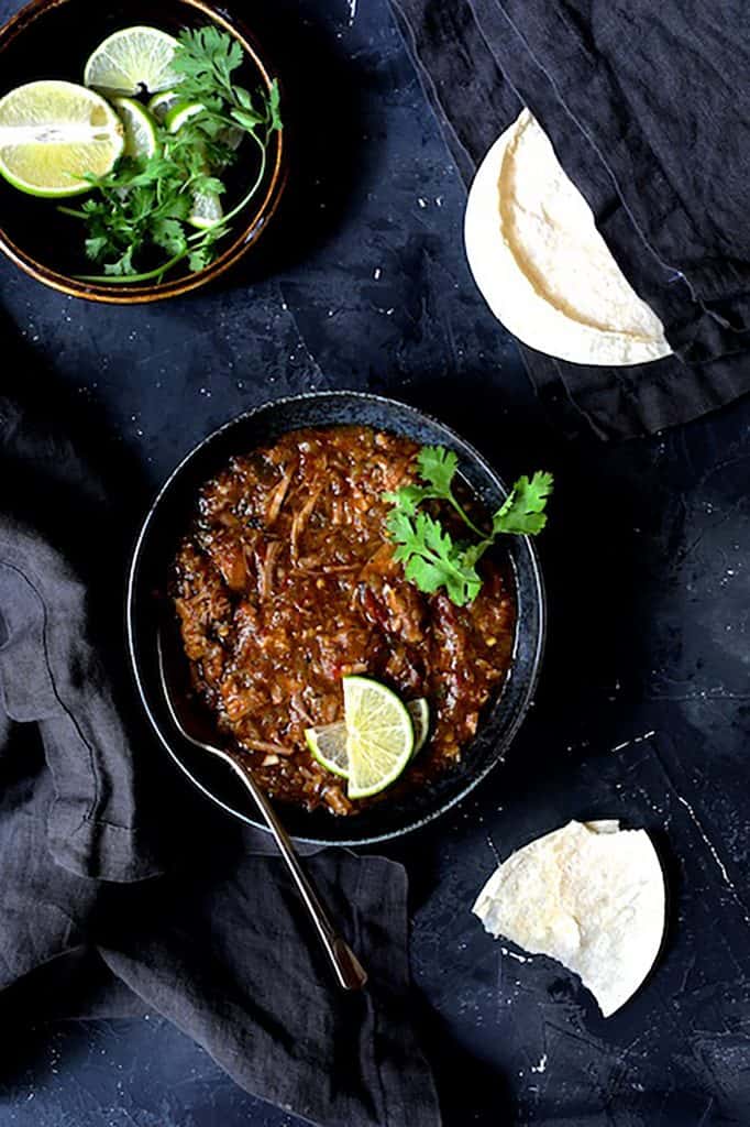 Photo of one serving of Slow Cooker Colorado Green Chili in black bowl on black background with lime wedges, cilantro sprigs and corn tortillas.