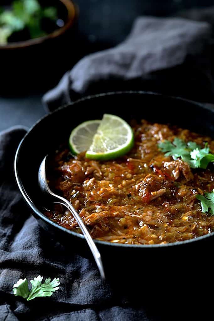 Photo of one serving of Slow Cooker Colorado Green Chili in black bowl on gray napkin garnished with cilantro sprigs and lime wedges.