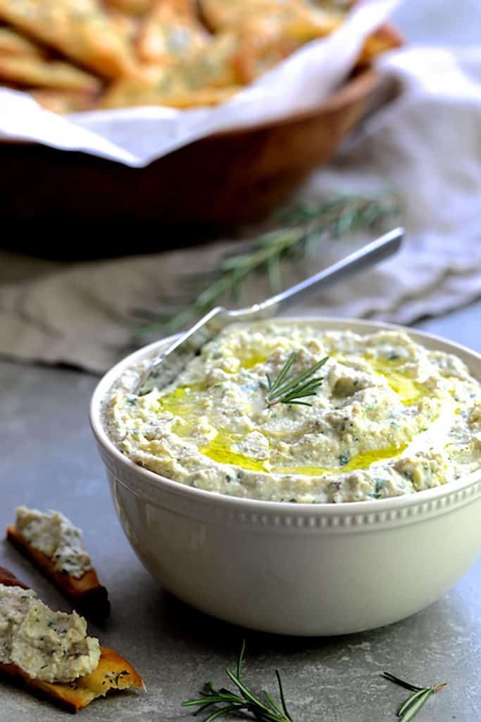 Photo of White Bean Artichoke Spread in white bowl with Rosemary Sea Salt Flatbread Chips in the background. 