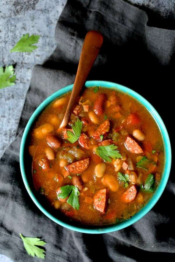 Photo of Slow Cooker Cajun 15 Bean Soup with Andouille Ham and Bacon in an aqua-colored bowl with copper spoon on gray napkin.