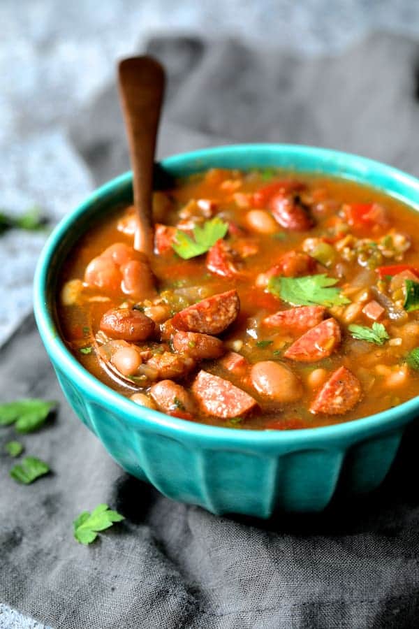 Photo of Slow Cooker Cajun 15 Bean Soup with Andouille Ham and Bacon in aqua-colored bowl with copper spoon on gray napkin garnished with parsley leaves.