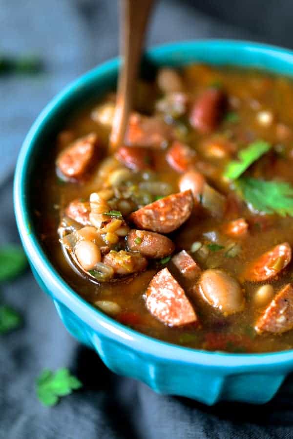 Close-up photo of Slow Cooker Cajun 15 Bean Soup with Andouille Ham and Bacon in aqua-colored bowl with copper spoon.