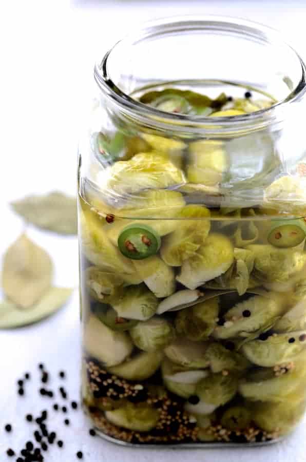 Photo of Quick Pickled Brussels Sprouts with Jalapeno in jar without the lid and black peppercorns and bay leaves on the table.
