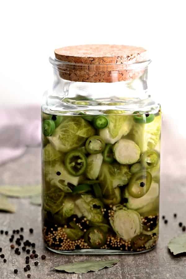 Photo of Quick Pickled Brussels Sprouts with Jalapeno in jar with lid on and bay leaves and black peppercorns on the gray table.