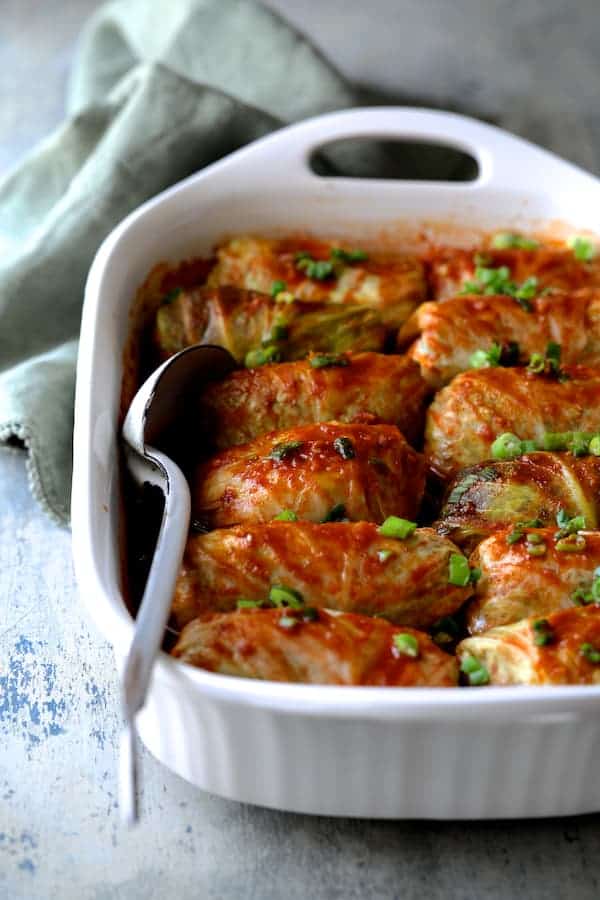 Photo of cooked Spicy Asian Pork Cabbage Rolls in white baking dish on blue background with green dish towel and white metal spoon.