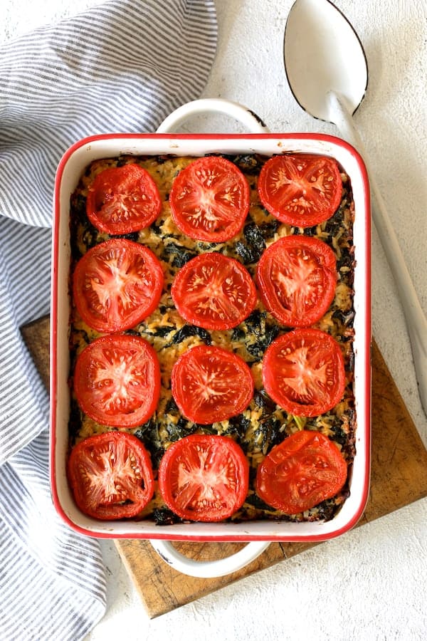 Photo of Brown Rice, Cheddar Kale Casserole in white pan with striped towel on wood trivet with white spoon.