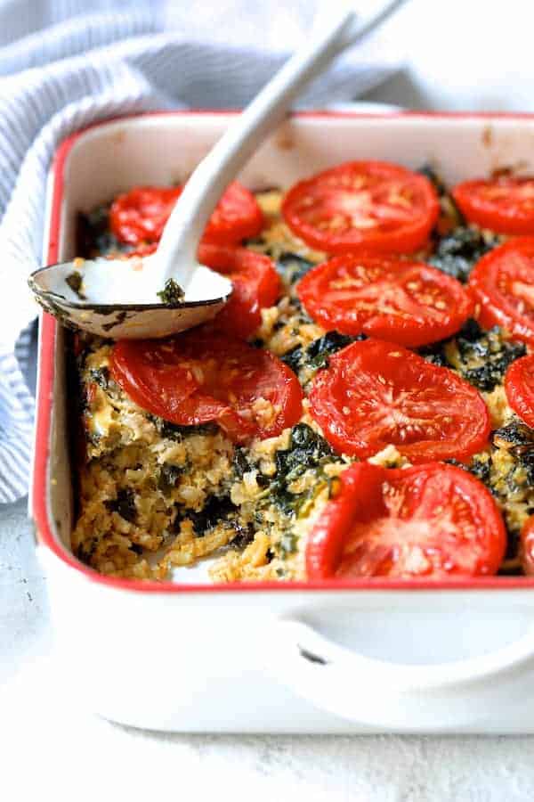 Photo of Brown Rice Cheddar Kale Casserole on white background with striped towel and white spoon with some served.