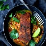 Indian Spiced Sea Bass with Braised Red Lentils in black bowl with fork, cilantro sprigs and lime wedges.