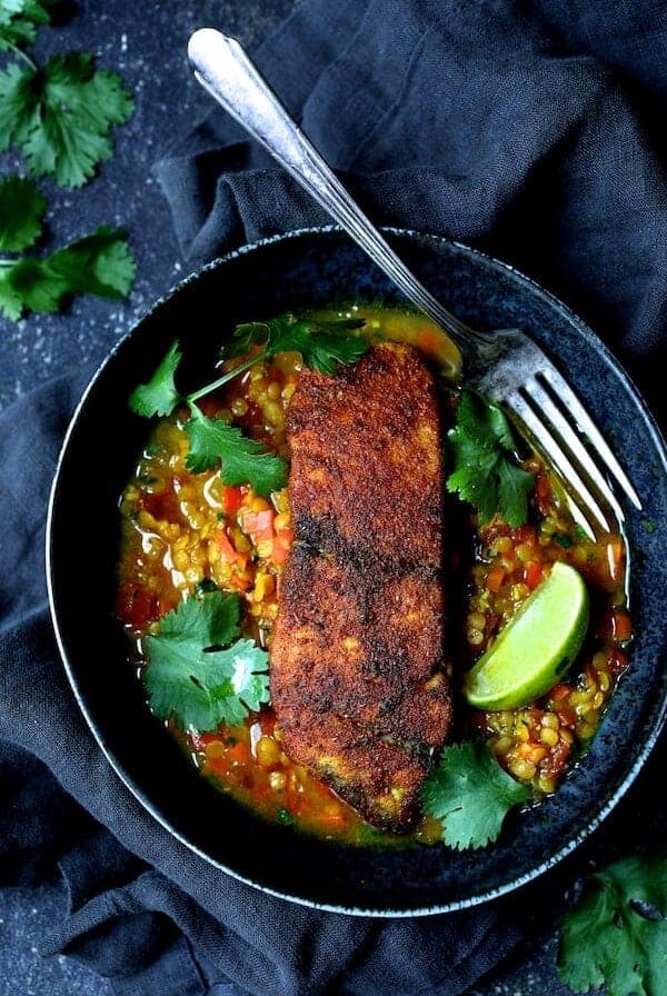 Indian Spiced Sea Bass with Braised Red Lentils in black bowl with fork, cilantro sprigs and lime wedges.
