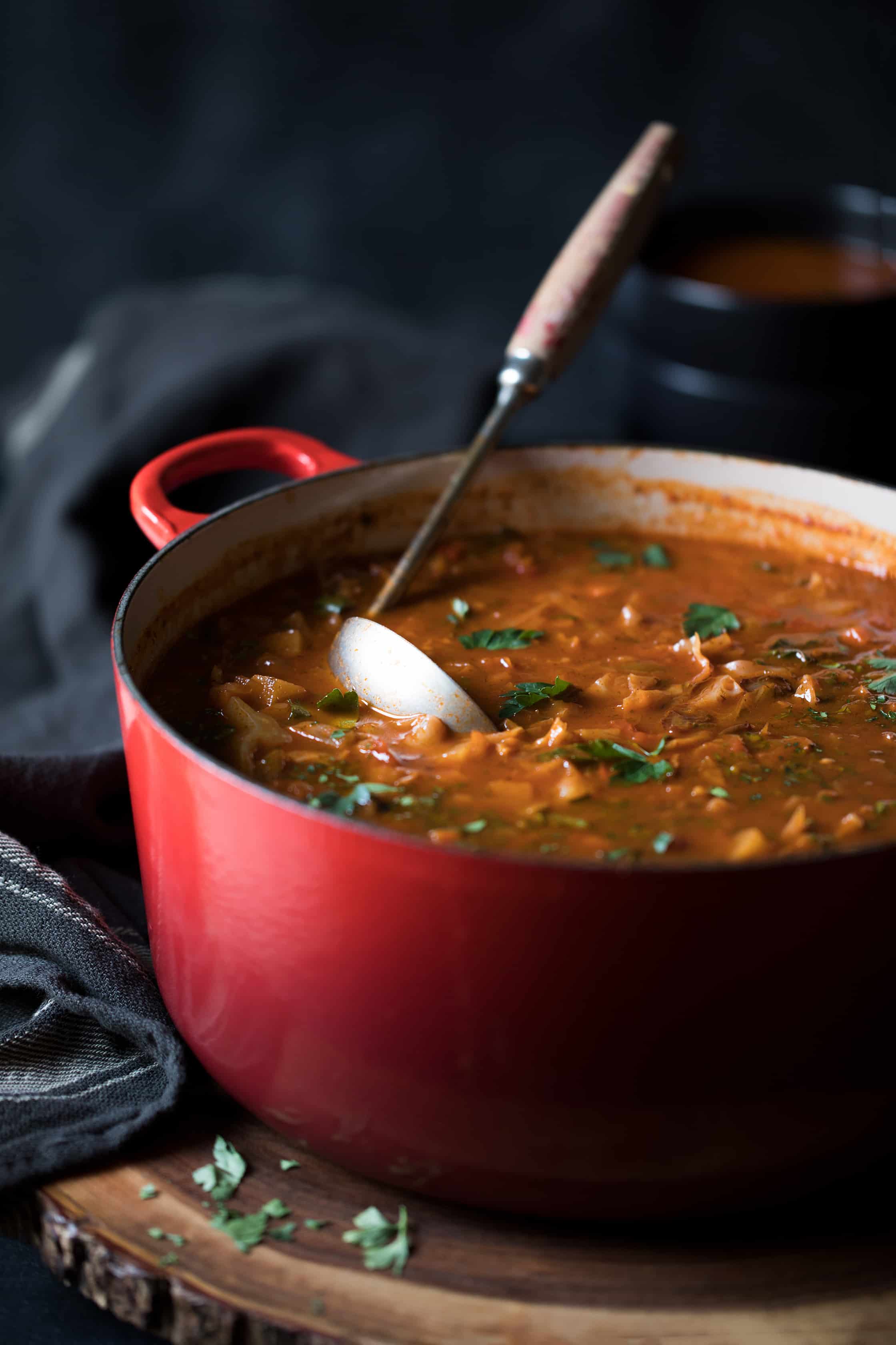 Photo of Rustic Cabbage and Root Vegetable Soup in red pot with ladle on wood trivet with striped towel.