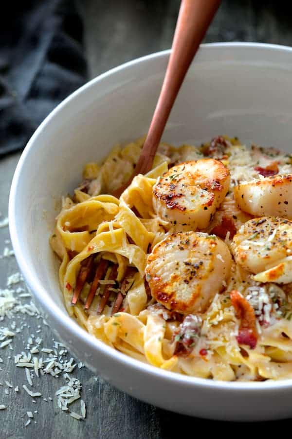 Close-up photo of Seared Sea Scallops with Spicy Bacon and Sun Dried Tomato Alfredo Sauce in white bowl with copper fork.