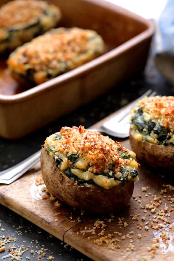 Photo of Spinach Parmesan Ranch Twice Baked Potatoes on small wooden cutting board with serving fork and clay baking dish in the background. 