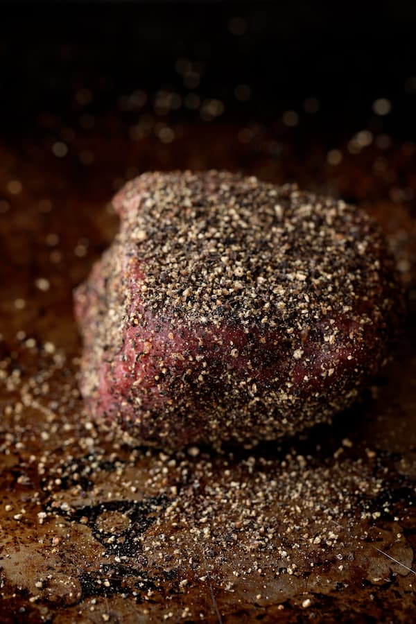 Photo of uncooked steak encrusted with black peppercorns.