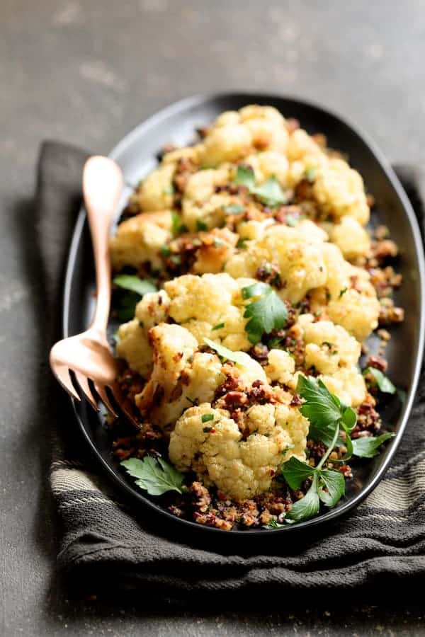 Photo of Roasted Cauliflower with Black Olive Pangrattato on black platter on gray striped towel and serving fork.