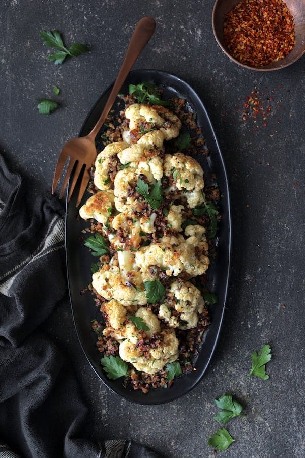 Photo of Roasted Cauliflower with Black Olive Pangrattato on black platter with copper serving fork garnished with fresh parsley.
