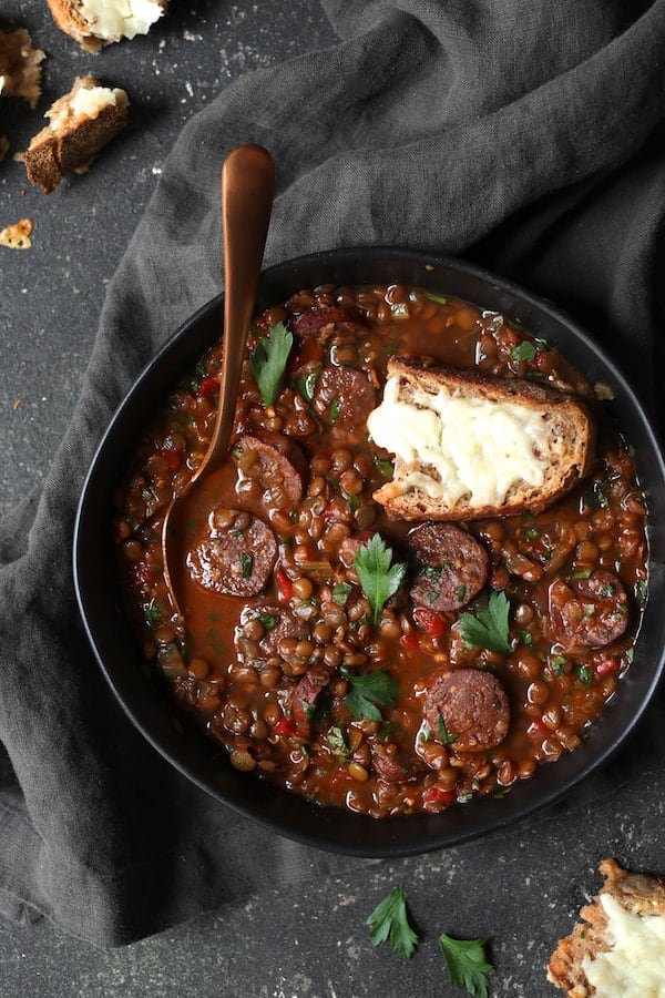 Photo of Soupy Spanish Lentils with Chorizo and Manchego Toast on gray background with gray towel.
