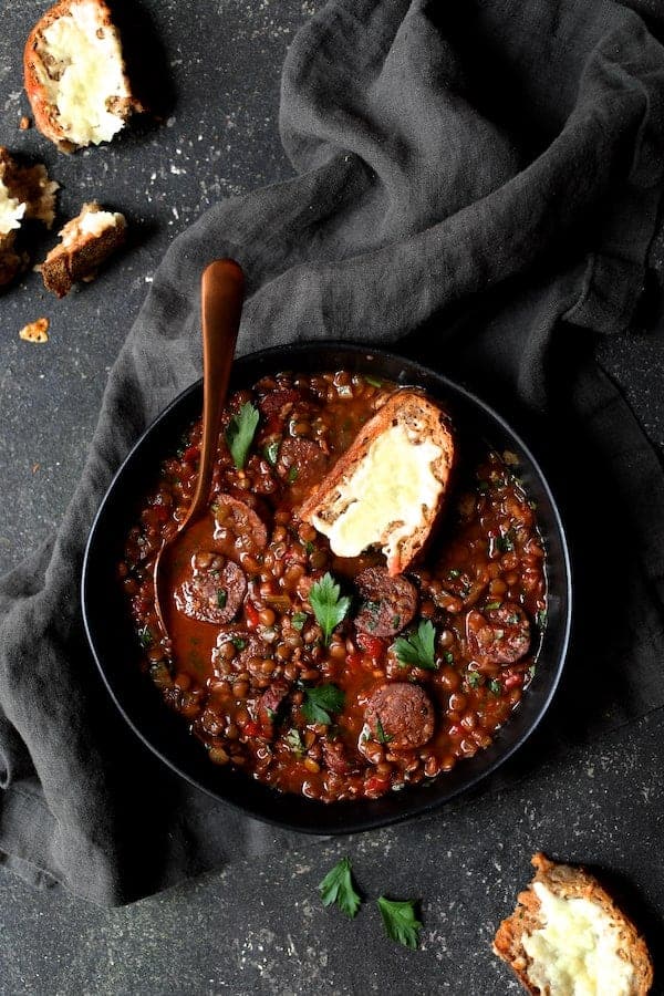 Photo of Soupy Spanish Lentils with Chorizo and Manchego Toast on dark gray background with gray napkin.