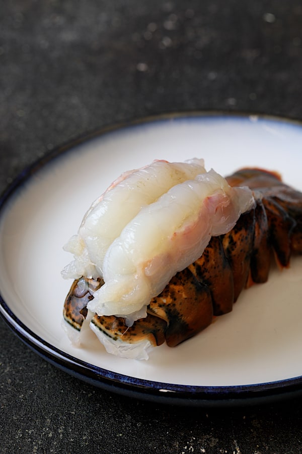 Photo of uncooked lobster tail with meat on top of the shell on white plate.