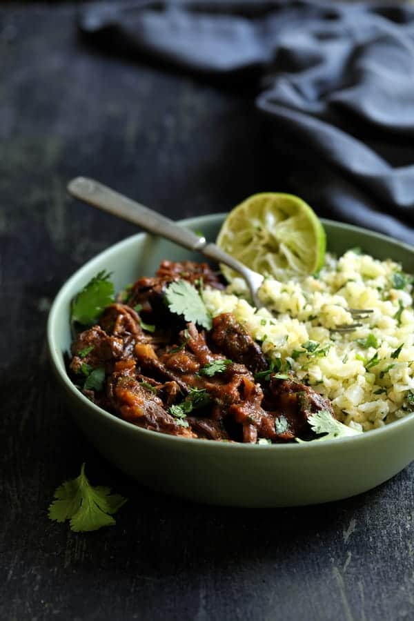 Photo of shredded Slow Cooker Beef Short Ribs Barbacoa with Cilantro Lime Cauliflower Rice in green serving bowl garnished with cilantro and lime with blue napkin in the background.