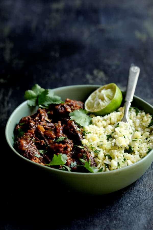 Photo of Slow Cooker Beef Short Ribs Barbacoa with Cilantro Lime Cauliflower Rice in green serving bowl garnished with cilantro and lime.