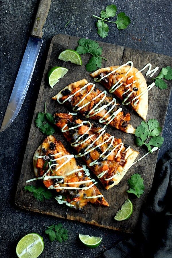 Photo of Chipotle Chicken Sweet Potato and Black Bean Flatbread Pizzas with Avocado Sour Cream cut into wedges on dark wood cutting board garnished with lime wedges and chopped cilantro.