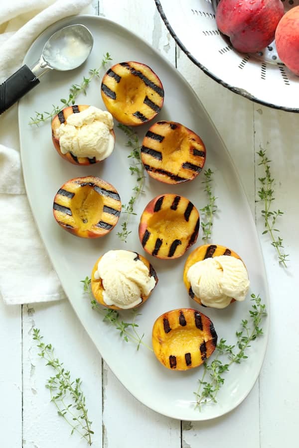 Photo of grilled peaches on white oval platter with ice cream added to three of the peaches.
