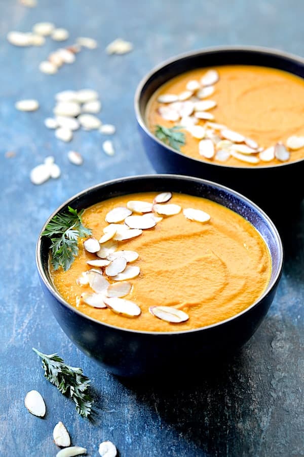 Photo of Spicy Indonesian Vegan Carrot Almond Soup in black bowls on blue background garnished with sliced almonds and carrot fronds.
