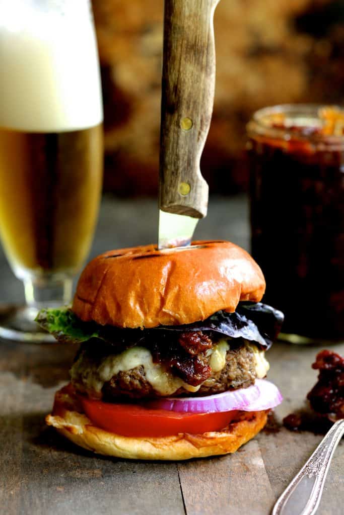 Photo of Grilled Burgers with Bacon Tomato Jam and Smoked Gouda with foamy beer in the background.