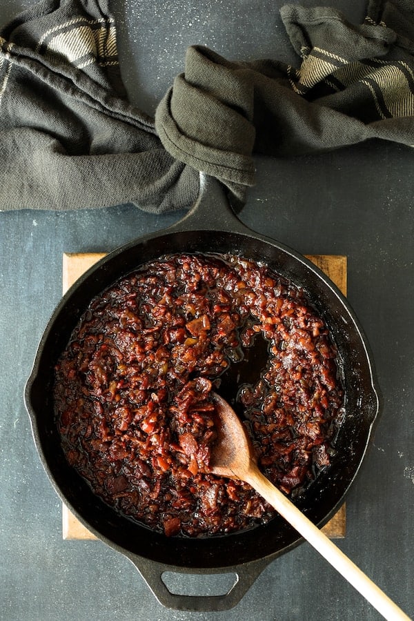 Photo of bacon jam in cast iron skillet on wood trivet with the handle of the skillet wrapped in gray striped kitchen towel.