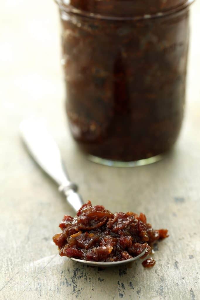 Close-up photo of bacon jam on spoon with glass jar of jam in the background.
