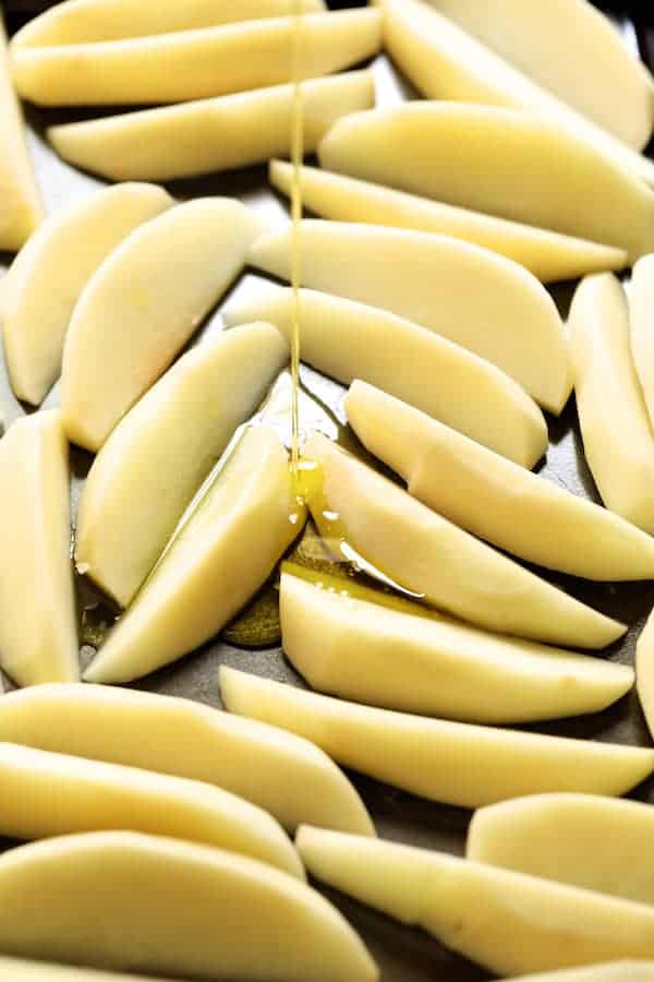 Photo of raw potatoes on baking sheet being drizzled with olive oil.
