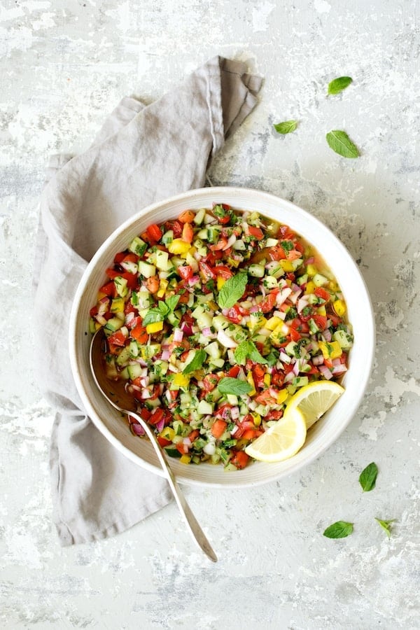 Photo of Middle Eastern Chopped Salad in white serving bowl on gray background with gray napkin garnished with lemon slices.