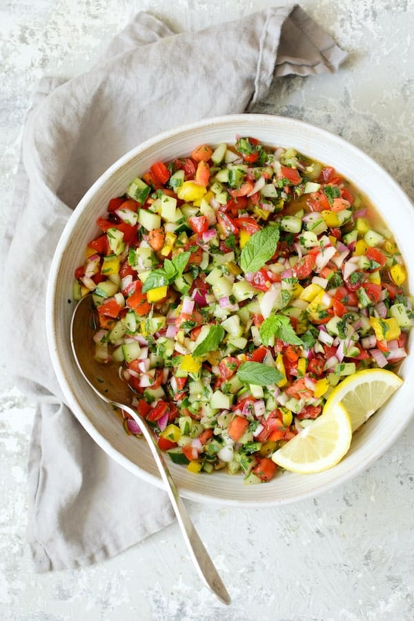 Photo of finished Middle Eastern Chopped Salad in white serving bowl on gray background with gray napkin garnished with lemon slices.