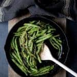 Charred green beans in cast iron skillet being stirred with wooden spoon.
