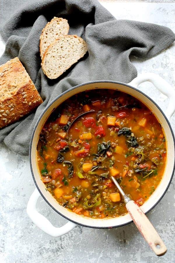 Photo of Spicy Kale Butternut Squash and Lentil Soup with Bacon in white enameled Dutch oven with whole grain bread on gray napkin.
