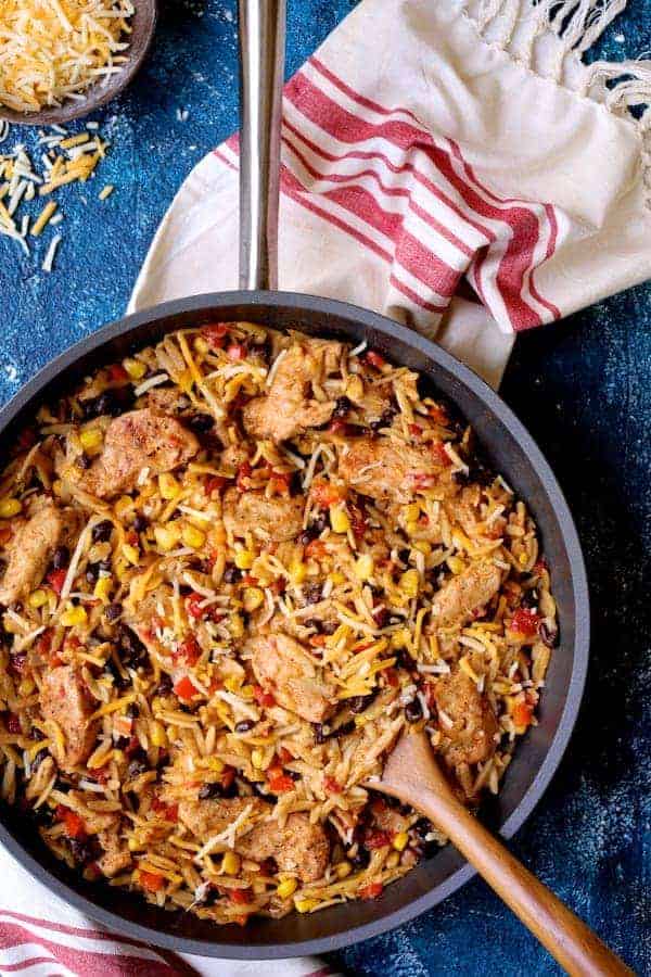 Photo of Southwestern Chicken Orzo and Black Bean Skillet with wooden spoon and red and white towel on blue background.