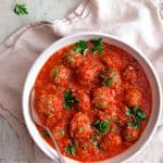 Spiced Meatballs with Tomato Sauce in white bowl with serving spoon on beige napkin.