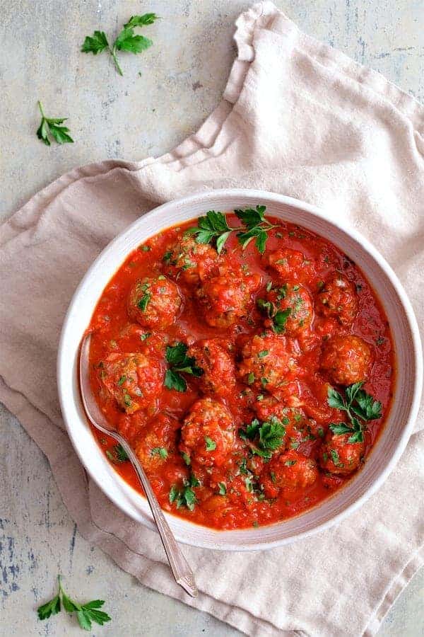Photo of Middle Eastern Meatballs in Tomato Sauce in white serving bowl on beige napkin garnished with parsley.