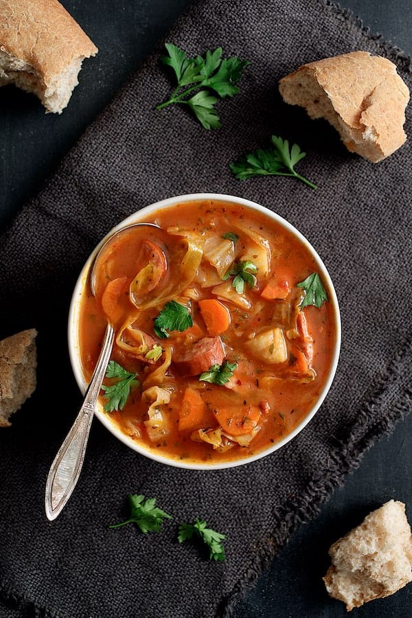 Photo of one serving of Old Fashioned Potato Cabbage and Kielbasa Soup in white bowl with gray napkin and bread.