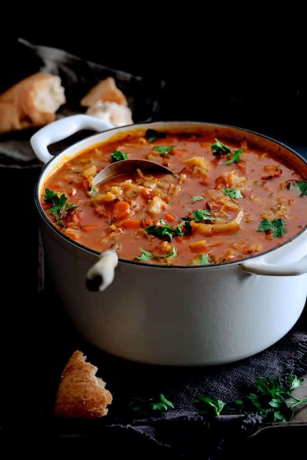 Photo of Old Fashioned Potato Cabbage and Kielbasa Soup in white enameled Dutch oven with ladle in the soup and bread scattered around.