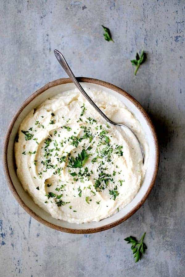 Photo of Cauliflower White Bean Puree in clay bowl with serving spoon garnished with parsley.