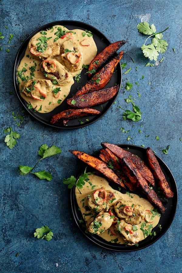 Photo of Spicy Tahini Pork Medallions with Harissa Roasted Sweet Potato Wedges on blue background garnished with cilantro sprigs.