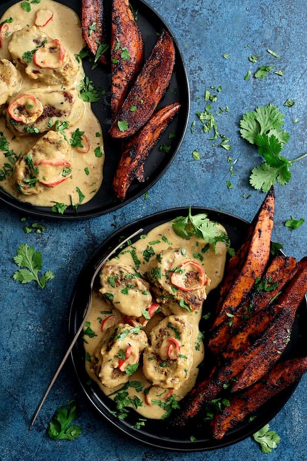 Photo of two servings of Spicy Tahini Pork Medallions with Harissa Roasted Sweet Potato Wedges on blue background garnished with cilantro.
