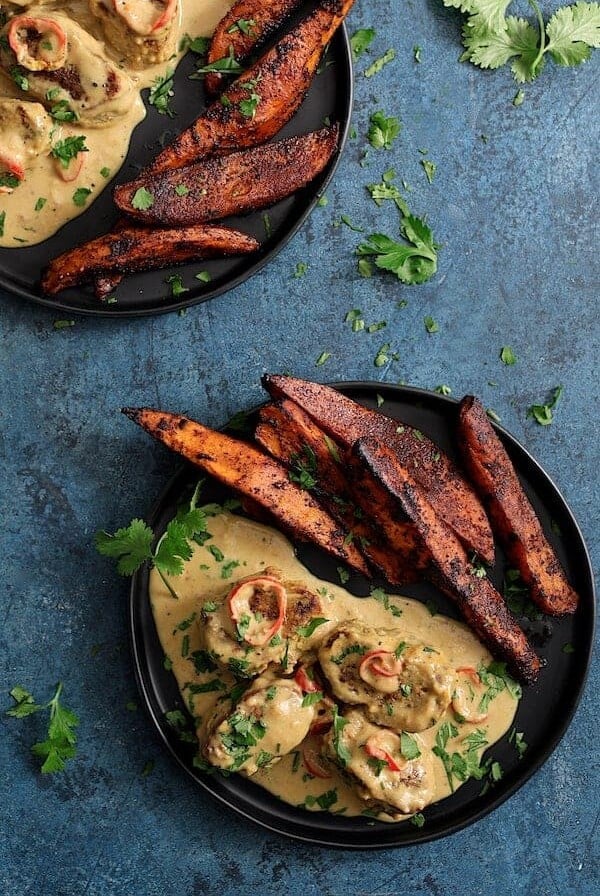 Two servings of Spicy Tahini Pork Medallions with Harissa Roasted Sweet Potato Wedges on black plates.