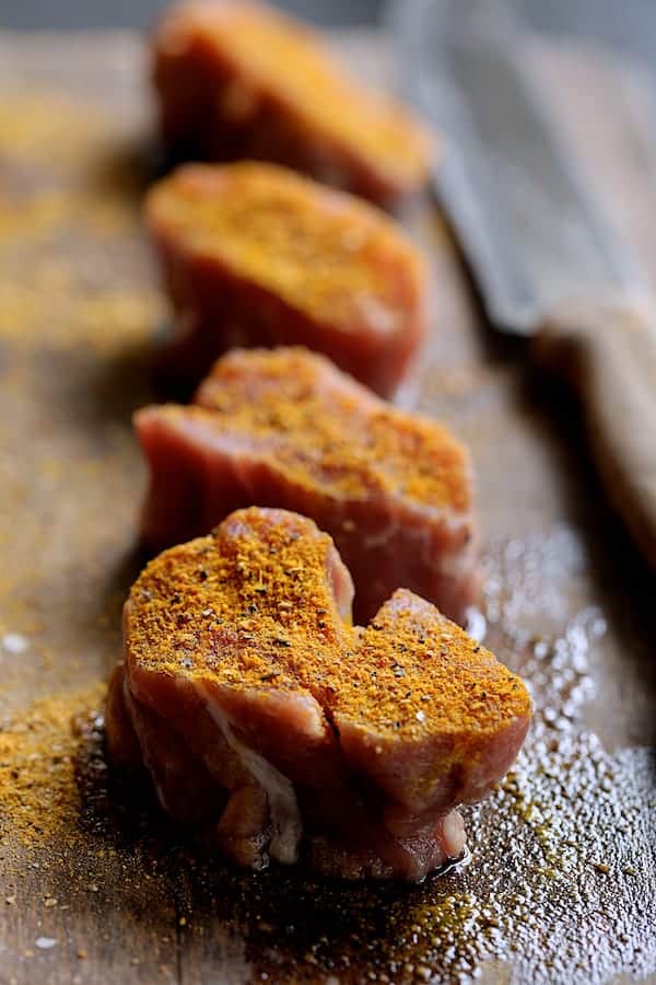 Photo of four uncooked pork medallions on wood cutting board sprinkled with spice rub.