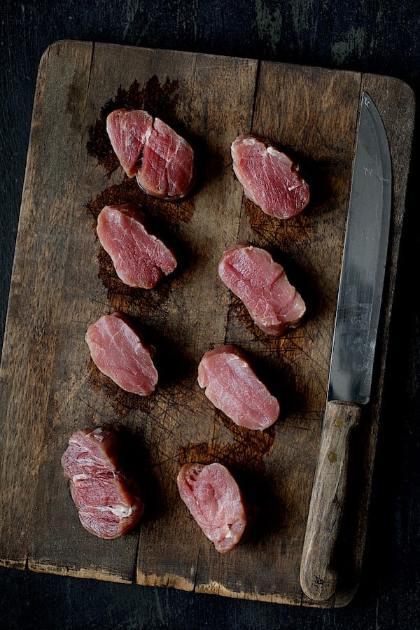 Photo of eight uncooked pork medallions on wood cutting board with knife.